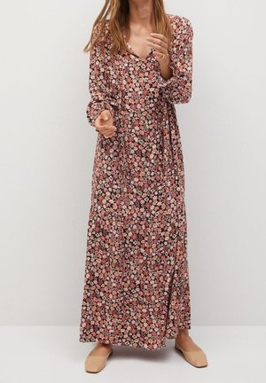 Vrouw draagt een lange bloemenjurk met roze, witte en oranje bloemen, gecombineerd met beige platte schoenen, staand tegen een effen achtergrond.
