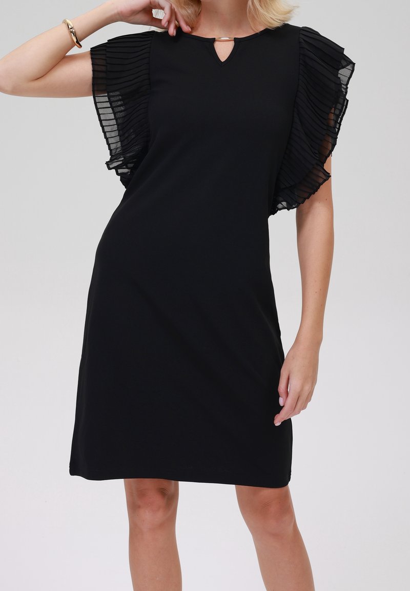 Femme portant une robe noire mi-longue avec des manches volantées plissées en voile transparent et un petit décolleté en forme de trou de serrure avec un fermoir doré.
