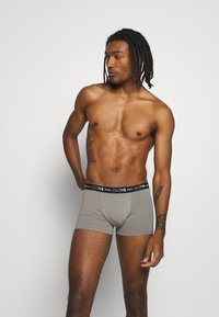 Graue Boxer-Shorts mit einem schwarzen markierten Bund, die eine enganliegende Passform und einen glatten Baumwollstoff aufweisen. Das Modell posiert im Profil vor einem neutralen Hintergrund.