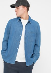 Tânăr îmbrăcat cu o cămașă albastră din denim cu nasturi peste un tricou alb, purtând o șapcă închisă la culoare și pantaloni închiși la culoare, privind spre stânga cu mâinile în buzunare.