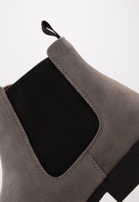 Botín de tobillo de ante gris con un panel lateral elástico negro, tirador en la parte superior y superficie texturizada. Suela de goma negra sólida.