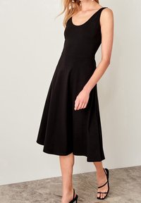 Femme portant une robe midi noire sans manches avec un décolleté rond et des sandales à talons noires à lanières, debout sur un sol en pierre.