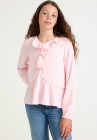 Ung pige med langt krøllet hår iført en lys pink bluse med flæser og blå jeans, stående mod en ensfarvet hvid baggrund.