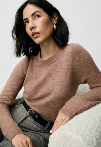 Hellbrauner Pullover aus Strickmaterial, mit langen Ärmeln und Rundhalsausschnitt, kombiniert mit karierten Hosen und einem schwarzen Gürtel.