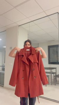 Person hält einen roten, doppelreihigen Mantel mit weitem Kragen drinnen hoch, vor einem Spiegel, der einen Raum mit Tischen und Stühlen reflektiert.