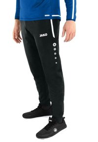Zwarte joggingbroek met wit JAKO-logo, voorzien van vijf zilveren knopachtige accenten aan de zijkant, gecombineerd met zwarte sportschoenen.
