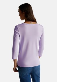 Donna con lunghi capelli castani, indossa una maglietta a maniche lunghe di un leggero colore viola e jeans blu, vista di spalle con un piccolo tatuaggio sul collo visibile.