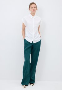 Witte button-up shirt met korte mouwen, gecombineerd met teal wijde broeken en puntige schoenen met een koeienprint.