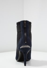 Bottine en suède bleu marine avec un talon aigu fin. Elle présente un panneau latéral élastique noir et une fermeture éclair à l'arrière, affichant une plaque de logo.