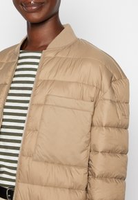 Béžová prošívaná péřová bunda se vzorem stehování, žebrovaným límcem a přední kapsou. Nasažena přes pruhované zeleno-bílé tričko.