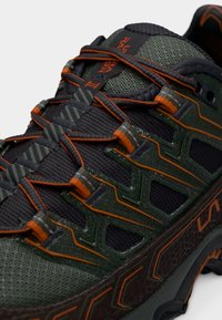 Παπούτσι για τρέξιμο σε μονοπάτι με πράσινο και μαύρο δίχτυνο επάνω μέρος, πορτοκαλί λεπτομέρειες, υφασμένες επικαλύψεις και ανθεκτική σόλα για κράτημα.