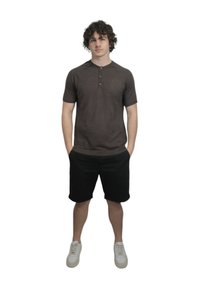 Maglietta henley corto a maniche corte di colore marrone scuro con patta a tre bottoni, abbinata a pantaloni corti neri e sneakers bianche, in piedi su uno sfondo bianco.