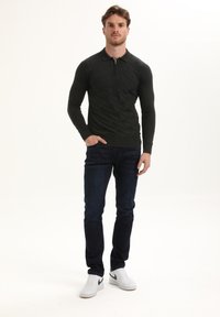 Vyras stovi su ranka kišenėje, dėvėdamas tamsiai raštuotą ilgomis rankovėmis polo marškinėlį, tamsias džinsus ir baltus sportbačius su juodu ženkleliu.