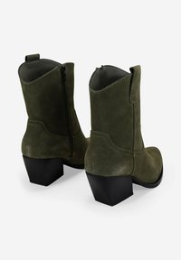 Un par de botines de ante verde oliva con cremalleras laterales, tiradores y tacones negros gruesos, mostrados desde la parte trasera sobre un fondo blanco.
