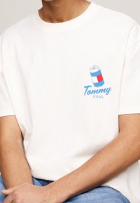 White cotton t-shirt featuring a blue and red graphic of a soda can. "Tommy Jeans" is written in cursive below the design.