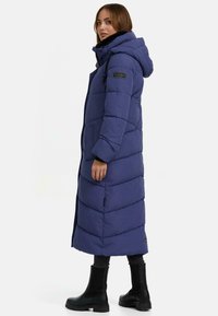 Cappotto lungo imbottito blu navy con design a righe orizzontali, cappuccio regolabile, tasche anteriori e dettagli in metallo nero. Finitura opaca, materiale caldo.