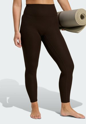 Brown ribbed leggings with a high waistband, featuring a smooth texture. The model holds a rolled yoga mat in neutral tones.