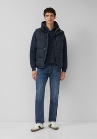 Granatowa puchowa kamizelka na ciemnoniebieskiej bluzie z kapturem o fakturowanej tkaninie, zestawiona z niebieskimi dżinsami i białymi tenisówkami z szarymi akcentami.