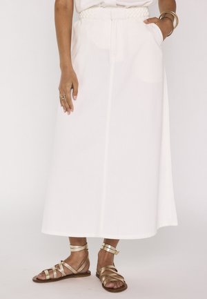 Femme portant une jupe midi blanche avec une taille élastique et des sandales plates dorées à brides, une main dans la poche et l'autre reposant à ses côtés.