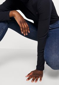Top negro de cuello vuelto con mangas largas, combinado con jeans de mezclilla azul oscuro. Primer plano de manos reposando sobre una superficie.
