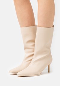 Beige nahkasaappaat nilkkoihin, joissa on kärki ja kapeat 5 cm korko. Tasainen pinta; varsi on puhdas ja minimalistinen muotoilu.