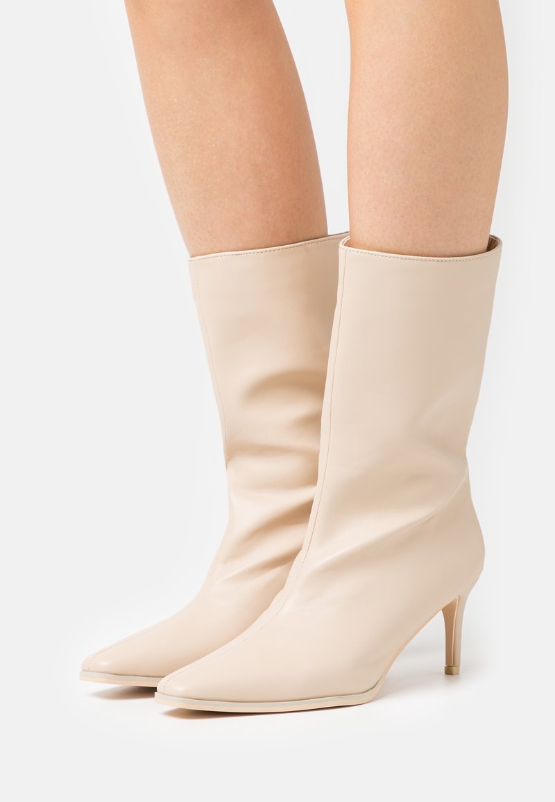 Beige nahkasaappaat nilkkoihin, joissa on kärki ja kapeat 5 cm korko. Tasainen pinta; varsi on puhdas ja minimalistinen muotoilu.