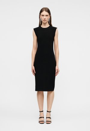 Nő ujjatlan, térdig érő fekete ruhában, fekete pántos magassarkúban és hosszú, lógó fülbevalókkal, egy egyszerű, világos háttér előtt állva.