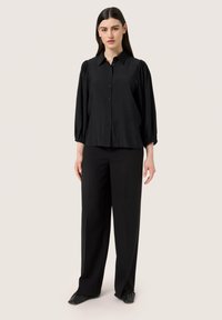 Blusa negra con cuello, con cierre de botones y mangas abullonadas, combinada con pantalones negros de pierna ancha y zapatos tipo slip-on, sobre un fondo neutro.
