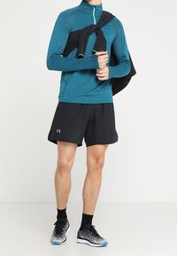 Teal lange mouwen sporttop, zwarte shorts en geprinte hardloopschoenen. Zwart kledingstuk over de schouder gehangen. Casual sportoutfit.