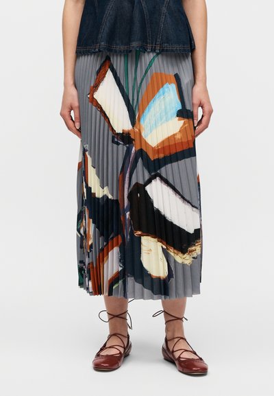 Jupe midi plissée avec un imprimé papillon en gris, orange et bleu. Portée avec des ballerines marron à lacets et un haut en denim foncé.