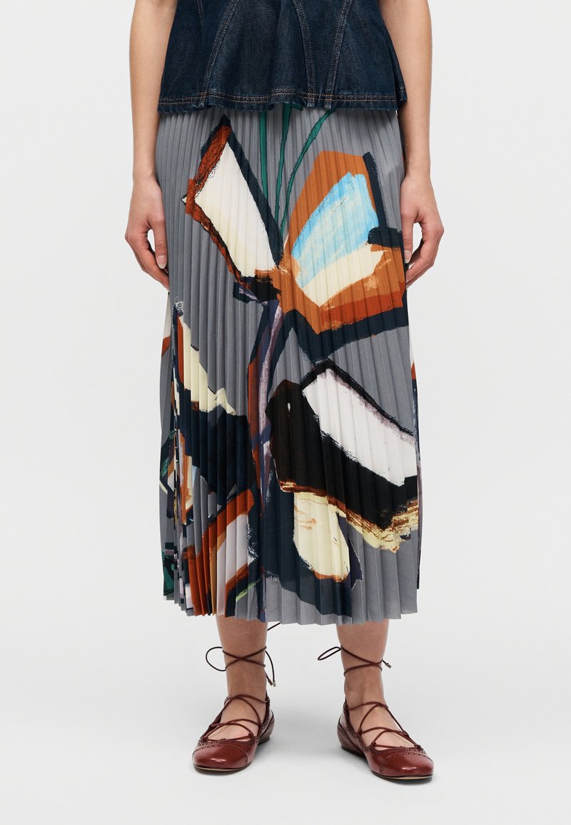 Jupe midi plissée avec un imprimé papillon en gris, orange et bleu. Portée avec des ballerines marron à lacets et un haut en denim foncé.