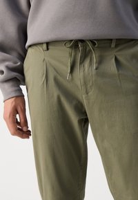 Pantaloni verde măsliniu cu o croială relaxată, dotati cu un sistem de strângere la talie și un design plisat. Construiți dintr-un material moale cu o textură netedă.