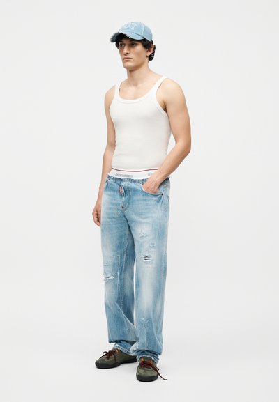 Νέος άνδρας που φοράει άσπρο αμάνικο, ανοιχτόχρωμα σκισμένα τζιν, μπλε καπέλο από τζιν και σκούρα αθλητικά παπούτσια, στέκεται με το ένα χέρι στην τσέπη.