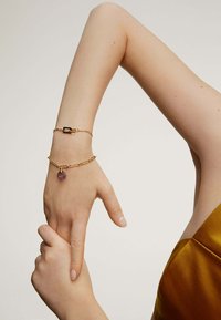 Pulseras doradas en una muñeca que presentan un cierre rectangular y una cadena con un charm de amatista, sobre un fondo neutro.