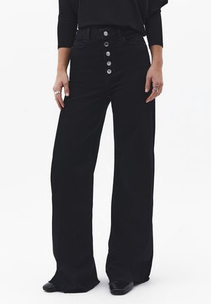 Person iført højtaljede, vidde ben sorte bukser med fem sølvknapper og sorte spidse sko, der står mod en hvid baggrund.