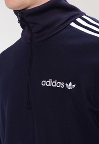 Sudadera navy con cremallera y cuello alto, logo de Adidas en blanco y tres franjas decorativas en los hombros, hecha de una suave tela texturizada.