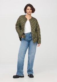 Giacca trapuntata verde oliva con bottoni a pressione, abbinata a una maglietta bianca e jeans ampi azzurri chiari. Scarpe nere con suola spessa.