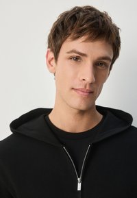 Hanorac negru cu fermoar și glugă cu șnur, purtat peste un tricou negru. Are un fermoar argintiu și o textură moale. Fața este parțial vizibilă.