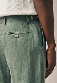 Hellgrüne Leinenhose mit strukturiertem Finish, ausgestattet mit einer Gesäßtasche mit Knopf und einem flachen Vorderdesign, gezeigt auf Taille.