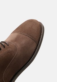 Scarpe da abito in suede marrone con punta arrotondata, dettagli di cucitura discreti e suola in gomma nera. I lacci sono scuri e presentano hardware minimale.