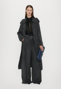 Grijze suède trenchcoat over een zwarte doorzichtige blouse, gecombineerd met wijde grijze broek en een textuur blauwe clutch met zilveren accenten.