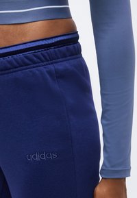 Άτομο που φοράει σκούρο μπλε παντελόνια Adidas και ανοιχτό μπλε μακρυμάνικη μπλούζα, εμφανίζοντας την κοιλιά και το δεξί χέρι προς τα κάτω.