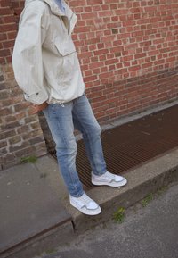 Giacca leggera bianca con tasca frontale, jeans azzurri, e scarpe da ginnastica bianche con dettagli grigi, su uno sfondo di muro di mattoni.