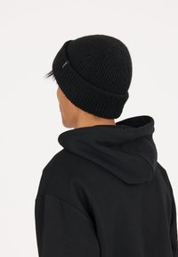 Svart stickad mössa med ribbat mönster, rundad topp och en liten varumärkesetikett, bärs över en svart huvtröja.