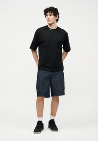 Bărbat tânăr cu păr creț, purtând un tricou negru și pantaloni scurți bleumarin cu dungi fine, stând cu mâinile la spate pe un fundal alb simplu.