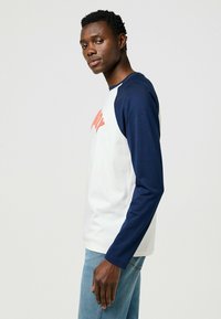 Langärmliges Raglan-T-Shirt in Weiß und Marineblau, das ein auffälliges oranges Logo auf der Vorderseite zeigt. Weicher Baumwollstoff mit körperbetontem Schnitt.