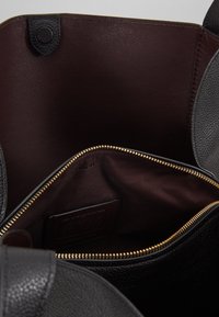 Sort læderhandbag med mørkebrunt interiør og en guld lynlås-lomme indeni, der viser fine tekstur- og syningsdetaljer.