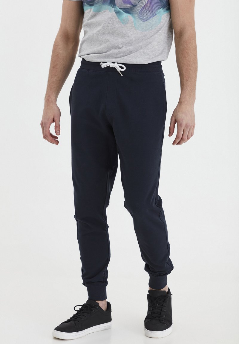 Navyblaue Sweatpants aus weichem Material mit elastischem Bund und weißem Kordelzug, schmal zulaufenden Beinen und gerippten Säumen. Schwarze Sneakers getragen.