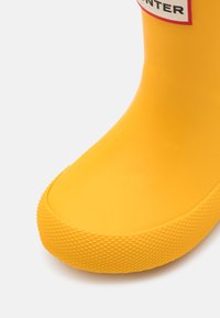 Vue de face d'une botte de pluie en caoutchouc jaune vif avec une semelle texturée et une étiquette partielle rouge et blanche près du haut.