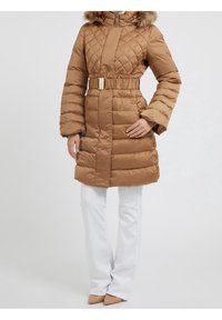 Abrigo acolchado marrón con capucha con borde de piel, cintura ceñida con cinturón y bolsillos laterales. Lleva puesto sobre pantalones blancos y tacones beige puntiagudos.